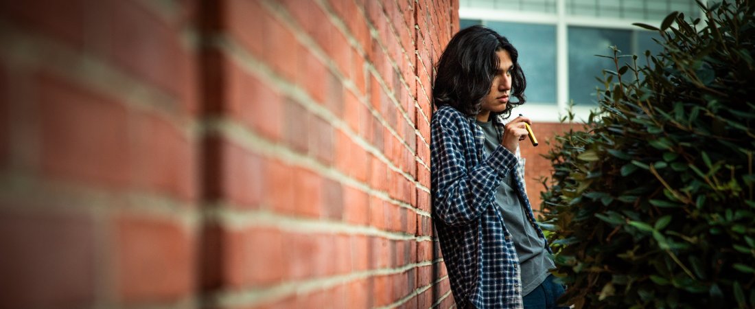 Un adolescente con pelo largo se apoya en una pared de ladrillos mientras sostiene un dispositivo para vapear. Este blog plantea los riesgos del vapeo para los adolescentes.