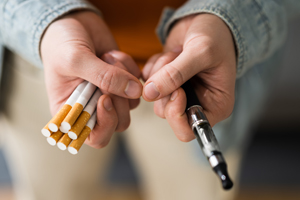 a pair of hands, holding a bundle of cigarettes in one and a vape pen in the other